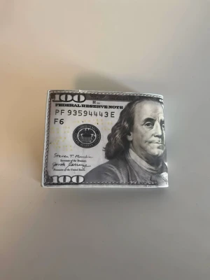 Plånbok med 100-dollar tryck - Säljer en unik plånbok i konstläder med tryck som liknar en amerikansk 100-dollarssedel. Plånboken har klassisk vikbar form och detaljer i svart, vitt och grönt med inslag av guld. Perfekt för dig som vill sticka ut med en rolig accessoar.