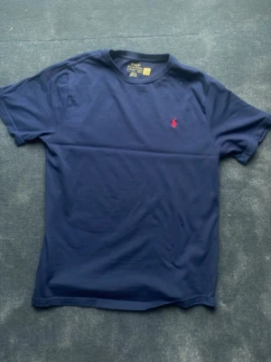 Marinblå t-shirt från Polo Ralph Lauren - Snygg marinblå t-shirt från Polo Ralph Lauren med klassisk passform och korta ärmar. T-shirten har en liten röd broderad logga på bröstet och är tillverkad i mjuk bomull. Perfekt för en stilren och avslappnad look.