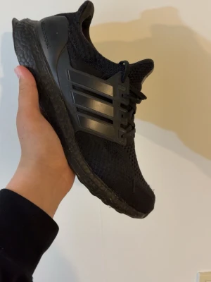 Svarta Adidas Ultraboost sneakers - Säljer ett par helsvarta Adidas Ultraboost sneakers med stickad ovandel och klassiska tre ränder på sidan. Skorna har en chunky Boost-sula för maxad komfort och en sportig vibe. Perfekta för dig som gillar streetstyle och vill ha något som funkar till allt.