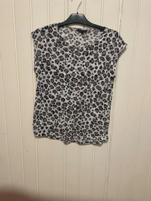 Leopardmönstrad topp från Alice Bizous - Snygg topp från Alice Bizous i storlek S med leopardmönster i svart, grått och ljusrosa på vit botten. Modellen har korta ärmar och rund halsringning. Perfekt för dig som gillar djurmönstrat och vill sticka ut lite extra.