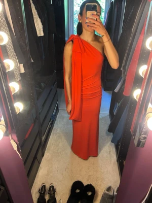 Röd oneshoulder långklänning - Snygg röd långklänning med oneshoulder-design och öppen rygg. Klänningen har en draperad detalj över axeln och smickrande passform. Korall/orange/röd i färgen. Säljer då den är lite kort på mig. 