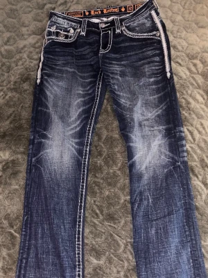 Rock Revival blå bootcut jeans - Snygga blå bootcut/straight jeans från Rock Revival med slitna detaljer och kontrastsömmar. Jeansen har dekorativa bakfickor med broderi och tydliga tvättade partier framtill och baktill. Perfekta för dig som gillar en edgy och trendig stil. Pris kan diskuteras