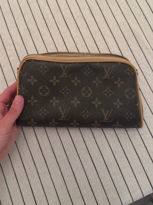  monogram väska  - Väska i klassiskt brunt monogrammönster med beige kanter och dragkedja. Själva dragaren från dragkedjan saknas. 