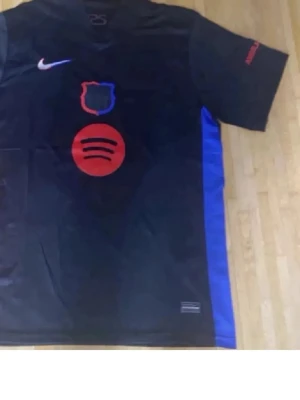 FC Barcelona svart matchtröja Nike - Svart FC Barcelona matchtröja från Nike med röd Spotify-logga på bröstet och klubbmärke i blått och rött. Blå och röda detaljer på sidorna och ärmarna. Kortärmad och tillverkad i lätt funktionsmaterial, perfekt för fotbollsträning eller match.