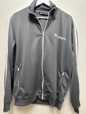 Grå track jacket - Snygg grå track jacket med vit dragkedja och vita detaljer längs ärmarna. Jackan har ståkrage, två fickor med dragkedja och logga på bröstet. Perfekt för en sportig och trendig look.