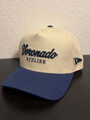Voronado Atelier Keps - Snygg keps i beige med blå skärm från Voronado Atelier. Broderad logga framtill och justerbar snapback baktill. Kepsen har en klassisk form och är tillverkad i bomullstyg. Perfekt för en cool och avslappnad stil.