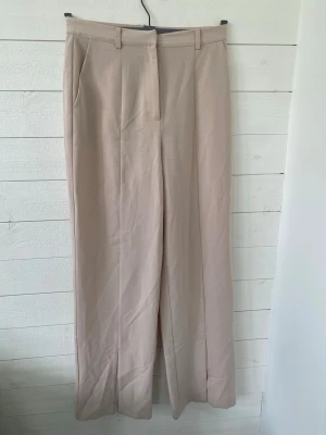 Beige vida kostymbyxor - Snygga beige kostymbyxor med vida ben och pressveck samt slits vid anklarn. Byxorna har hällor för skärp, snedställda fickor fram. Perfekta för en stilren och trendig look.