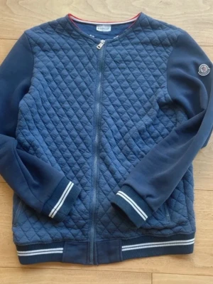 Vintage Blå quiltad jacka från Moncler - Snygg blå quiltad vintage jacka från Moncler med diamantmönstrad insida och kontrastkant i vitt och rött. Jackan har dragkedja och ribbade muddar. Perfekt för dig som vill ha en stilren och exklusiv look.