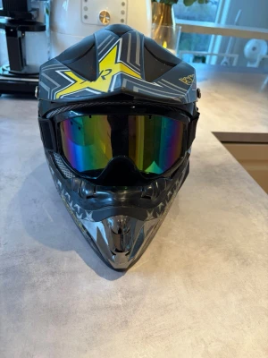 Rockstar crosshjälm med glasögon - Säljer en svart och grå crosshjälm med coola Rockstar Energy Drink-motiv och stora gula stjärnor. Hjälmen har matchande spegelglasögon med regnbågsfärgad lins. Perfekt för dig som gillar motocross eller enduro och vill sticka ut.