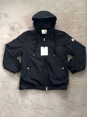 Svart vindjacka från Moncler med huva - Snygg svart vindjacka från Moncler med dragkedja, huva med dragsko och två fickor framtill. Jackan har Moncler-logga på ärmen och diskreta vita och röda detaljer på huvan. Perfekt för blåsiga dagar och enkel att matcha med olika outfits. XL (170-175cm) (70-80kg)