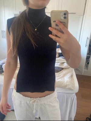 Svart ribbad polotopp med slits - Snygg svart ribbad polotopp med höga slitsar i sidorna. Toppen är ärmlös och har en tight passform som framhäver figuren. Perfekt för dig som gillar en stilren och trendig look.