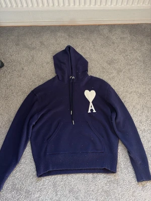 Mörkblå hoodie från Ami Paris - Säljer en mörkblå hoodie från Ami Paris med stor vit hjärtlogga och bokstaven A på bröstet. Hoodien har dragsko i huvan, ribbade muddar och en klassisk känguruficka framtill. Materialet är stickat och känns riktigt mjukt och skönt. 