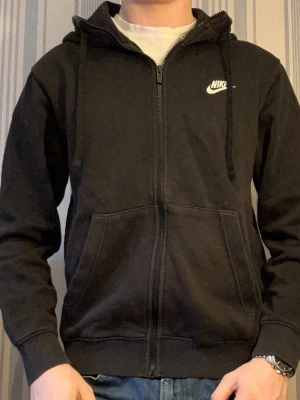 Svart zip-hoodie från Nike - Svart hoodie från Nike med dragkedja framtill, justerbar huva med snören och två stora fickor. Klassisk Nike-logga i vitt på bröstet. Tillverkad i mjukt bomullsmaterial som är skönt att ha på sig. Perfekt för en avslappnad och sportig stil.