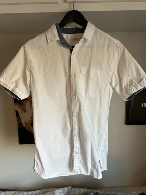Vit kortärmad skjorta  - Vit skjorta med korta ärmar och klassisk krage. Skjortan har en bröstficka och kontrastfärgad insida på kragen och ärmsluten. Perfekt för en clean och stilren look