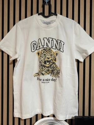 Vit GANNI t-shirt med leopardtryck - Vit t-shirt från GANNI med ett stort tryck av en leopardunge och texten 'Have a nice day!'. Klassisk passform med rund hals och korta ärmar. Perfekt för dig som gillar coola statement-plagg och djurmotiv.