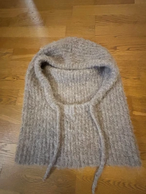beige grå stickad balaclava med snören - Mysig grå/beige stickad balaclava i mjukt garn med ribbat mönster och knytband framtill. Perfekt för kalla dagar när du vill hålla både huvud och hals varm. Enkel och stilren design som passar till det mesta.