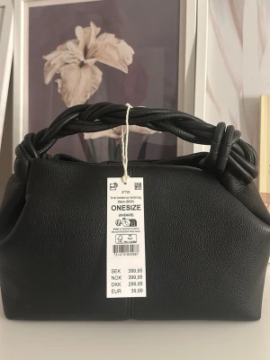 Svart handväska från Gina Tricot - Väskan är helt ny och oanvände och är perfekt för dig som vill ha en väska att gå runt med på stan! Trendig svart handväska från Gina Tricot med flätat handtag och stilren design. Väskan är tillverkad i syntetmaterial och har en mjuk, strukturerad yta. Perfekt accessoar för att lyfta din outfit.