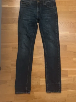 Mörkblå jeans från Nudie Jeans - Säljer ett par mörkblå jeans från Nudie Jeans med klassisk femficksdesign och snygga kontrastsömmar. Jeansen har rak passform och är tillverkade i denim med en cool tvätt och broderad bakficka. Perfekt för dig som gillar stilrena och tidlösa jeans. Storlek 28/32