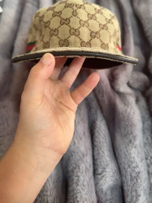 Gucci keps med GG-mönster - Snygg keps från Gucci i beige canvas med klassiskt GG-monogram över hela ytan. Kepsen har brunt innertyg, böjd skärm och detaljer i rött och grönt på sidorna. Perfekt accessoar för att lyfta din streetwear-look.