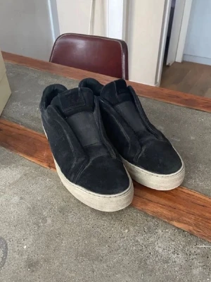 Svarta slip-on sneakers i mocka - Stilrena svarta slip-on sneakers med ovandel i mocka och tjock vit sula. Skorna har minimalistisk design utan snörning och en klassisk låg profil som passar till många olika outfits.