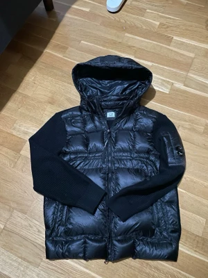 Svart cardigan jacka storlek L - Snygg svart cardigan jacka från C.P. Company med huva och stickade ärmar. Jackan har dragkedja framtill och en ficka med logga på ena ärmen. Perfekt för kalla dagar och riktigt trendig look.