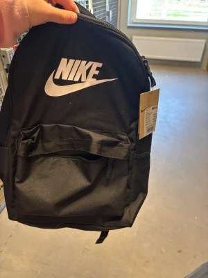 Svart ryggsäck från Nike - Snygg svart ryggsäck från Nike med stort vitt logotryck på framsidan. Ryggsäcken har ett stort huvudfack med dragkedja och en praktisk ytterficka framtill. Tillverkad i slitstarkt tyg och har justerbara axelband för bästa komfort. Oanvänd med prislapp kvar 