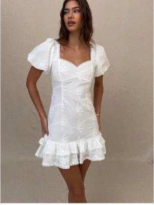 Vit klänning med volang,puffärmar och rosett - Söt vit kort klänning med puffärmar, volangkant och broderade blommor. Klänningen har öppen rygg med en stor rosett och dekorativa spetsdetaljer längs halsringningen. Klänningen är aldrig andvända och har prislappen kvar.