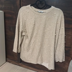 Beige tröja med pärlor från Zara - Snygg beige tröja från Zara med dekorativa pärlor framtill. Tröjan har rund halsringning, tre kvarts ärmar och en avslappnad passform. Perfekt för dig som gillar en stilren men ändå trendig look. Använd 2-3 gånger, gott som ny!