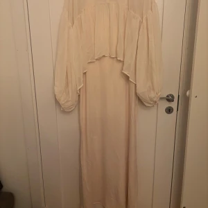 Lång ljus klänning med volangärmar - Elegant långklänning i ljus beige med voluminösa, transparenta långärmar och volangdetaljer upptill. Tröjan har en hög krage och är tillverkad i ett tunt, flowy material som ger en romantisk känsla. Klänningen är en lång tajt skims klänning