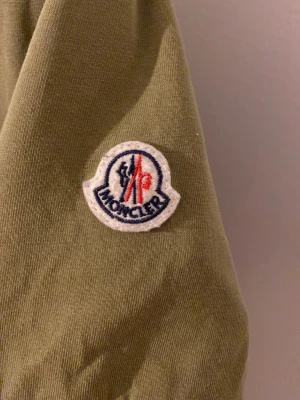 Olivgrön Moncler t-shirt i bomull - Snygg olivgrön t-shirt från Moncler med klassisk logotyp på ärmen och diskreta ränder i rött, vitt och blått vid ärmsluten. Tillverkad i mjuk bomull och har en normal passform. Perfekt för en stilren och avslappnad look.