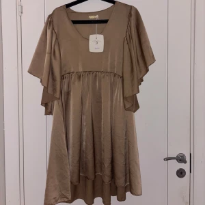 Beige omlottklänning med volang - Snygg beige klänning med v-ringning och volangärmar. Klänningen har omlottkänsla och ett lätt glansigt material som ger en lyxig look. Perfekt för dig som vill sticka ut med en elegant och trendig stil. Prislapp kvar