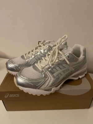 Vita och silver Asics sneakers - Snygga vita och silverfärgade sneakers från Asics med mesh- och syntetmaterial. Skorna har klassisk låg profil, vita snören och tydliga Asics-loggor på sidorna. Perfekta för dig som gillar sportig och trendig stil.