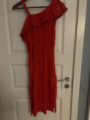 Röd långklänning med volang  - Säljer en röd långklänning från Lindex med volangdetaljer vid axlarna och en axelbandslös design på ena sidan. Klänningen har en smal passform och är tillverkad i mjuk bomull. Perfekt för dig som gillar färgstarka och trendiga plagg.
