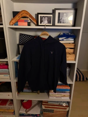 Mörkblå Cashmere hoodie från GANT - Snygg mörkblå hoodie från GANT med klassisk huva och dragsko. Tröjan har en broderad GANT-logga på bröstet och en stor magficka framtill. Tillverkad i mjukt material som känns skönt mot huden. Perfekt för en avslappnad och stilren look.