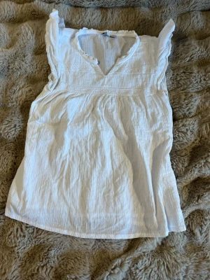 Vit ärmlös blus med volangdetaljer - Söt vit blus i tunn, krinklad bomull med spetsdetaljer och volangkant vid axlarna. Djup v-ringning med liten volang runt. Perfekt att matcha med jeans eller kjol för en fräsch look.