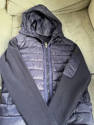 Mörkblå ”monclear” cardigan  - Snygg mörkblå pufferjacka med stickade ärmar och vadderad kropp. Jackan har en praktisk huva, dragkedja framtill och en ficka med dragkedja på ärmen. Perfekt för våriga dagar och ger en skön look vid intresse Ede bara att skriva