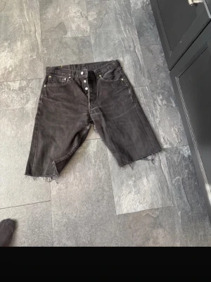 Svarta jeansshorts från Levi's - Säljer ett par svarta jeansshorts från Levi's med råa, avklippta benslut och klassisk femficksdesign. Shortsen har knappgylf och detaljer i silver. Perfekta för dig som gillar en avslappnad och edgy stil.