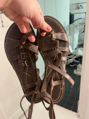 Bruna gladiatorsandaler med remmar - Säljer ett par bruna gladiatorsandaler med flera smala remmar och spännen. Sandalerna har dekorativa sömmar i kontrastfärg och långa snören som kan knytas runt ankeln. Tillverkade i konstläder för en cool och avslappnad stil.