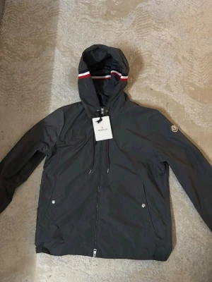 Moncler windbreaker - Snygg mörkgrå vindjacka från Moncler med huva och dragkedja framtill. Jackan har vita och röda ränder på huvan samt Moncler-logga på ärmen. Två fickor med tryckknappar och justerbar huva. Tillverkad i lätt, vattenavvisande material.