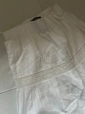 Vit långkjol med spetsdetaljer - Snygg vit långkjol med breda spetsdetaljer och volang. Kjolen har en luftig och flowy passform, perfekt för varma dagar. Tillverkad i ett lätt bomullstyg som känns skönt mot huden.