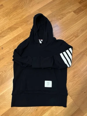 Thom Browne Hoodie - Svart hoodie från Thom Browne med vit patch framtill och fyra vita ränder på ena ärmen. Klassisk huva och känguruficka. Perfekt för dig som gillar stilrena och exklusiva plagg med en sportig touch.