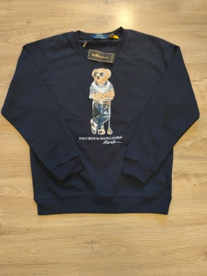 Mörkblå Polo Bear sweatshirt - Mörkblå sweatshirt från Ralph Lauren med ikoniska Polo Bear-trycket framtill. Tröjan har rund halsringning, långa ärmar och ribbade muddar vid ärmslut och nederkant. Perfekt för en avslappnad och trendig stil. Det står att den är Large men den är i storleken Small