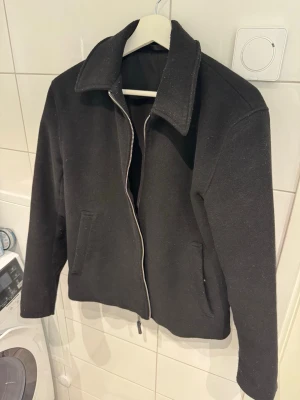 Svart ulljacka med dragkedja - Stilren svart jacka i ullmaterial med rak passform och bred krage. Jackan har två snedställda fickor framtill och stängs med en silverfärgad dragkedja. Perfekt för kyligare dagar och enkel att matcha med olika outfits.