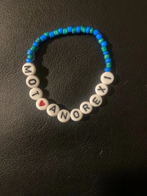 Blått pärlarmband med text - Ett handgjort armband med blå och turkosa pärlor samt vita bokstavspärlor som stavar 'MOT ANOREXI' med ett rött hjärta. Armbandet är elastiskt och har en ungdomlig, personlig stil. Perfekt för dig som vill bära ett statement.