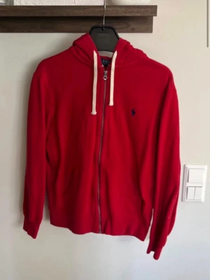 Röd hoodie från Polo Ralph Lauren - Röd hoodie med dragkedja från Polo Ralph Lauren. Klassisk design med vit dragsko i huvan och den ikoniska blå loggan broderad på bröstet. Mjuk bomullskänsla och ribbade muddar vid ärmslut och nederkant. Perfekt för en avslappnad stil.