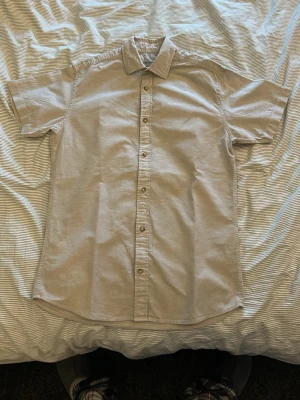 Beige kortärmad skjorta med knappar - Snygg beige skjorta med korta ärmar och klassisk krage. Skjortan har knappar hela vägen framtill och är tillverkad i ett lätt bomullstyg som passar perfekt till varmare dagar. Enkel och stilren design som är lätt att matcha.