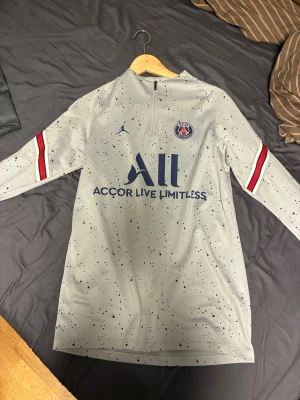 PSG träningströja grå Jordan - Grå långärmad träningströja från PSG x Jordan med mörkblå och röda detaljer på ärmarna. Tröjan har ett stänkmönster i mörkblått, halv dragkedja och PSG-logga på bröstet. Perfekt för fotbollsträning eller sportiga outfits.