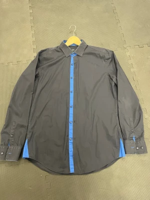Hugo Boss skjorta  - Stilren svart skjorta med blå knappslå och blå detaljer vid ärmslut och sidor. Klassisk krage och långärmad modell. Perfekt för dig som vill ha en skjorta med en twist. Tillverkad i mjuk bomull för skön känsla.