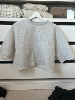 Grå croppad topp från Gina Tricot - Säljer en ljusgrå croppad topp från Gina Tricot. Toppen har rund halsringning, breda korta ärmar och är gjord i mjuk bomullsblandning. Perfekt till jeans eller kjol för en avslappnad look.