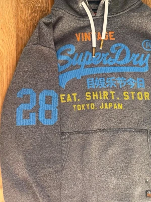 Superdry hoodie  - Snygg grå hoodie från Superdry i väldigt bra skick, finns en liten fläck vid fickan men inte alls så synlig. Skriv om du har frågor